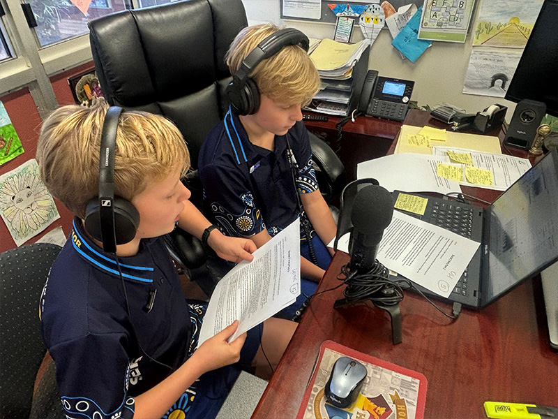 Two students at a desk with headphones on in front of a microphone and laptop.