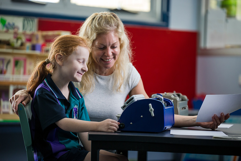 An adult and a student at a desk in a classroom setting, engaged in an activity together.